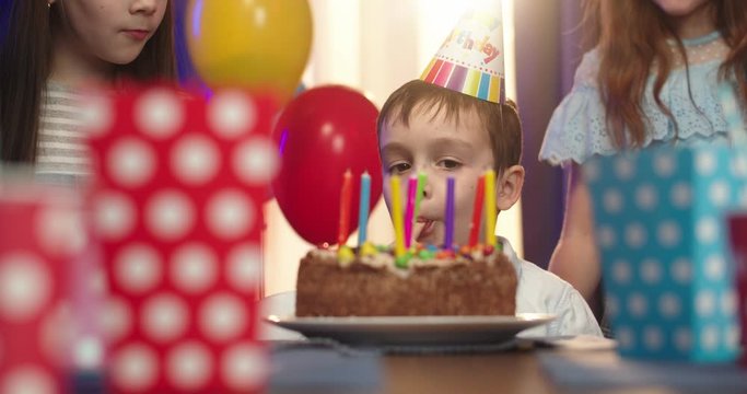 Funny Caucasian little boy in birthday conus sitting in front of the cake with candles and licking a cream from the top. Close up.
