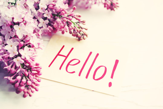 Text HELLO on a white card and lilac flowers on a white wooden table surface, closeup, selective focus.  Romantic background.