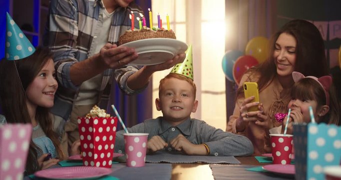 Caucasian cute birthday boy in conus sitting at the table with friends while father or older brother coming and bringing birthday cake.