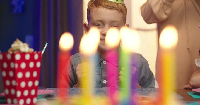 Portrait shot of the cute Caucasian small boy in the birthday conus looking at the cake with candles and waiting to blow them.