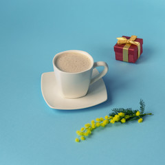 Cup of coffee, gift in a box and mimosa flower on blue background. Valentines day, birthday or Womens day concept.