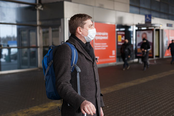 Young European man in gray coat, protective disposable medical mask in airport. Afraid of dangerous N-CoV 2019 influenza coronavirus mutated and spreading in China. Blue backpack, suitcase on wheels