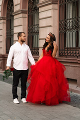 Fototapeta premium Beautiful romantic couple. Attractive young woman in red dress and crown with handsome man in white shirt with red rose walking on the street.Happy Saint Valentine's Day. Pregnant and wedding concept.