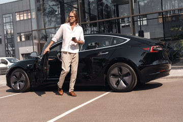 Fototapeta na wymiar Mode of Transport. Man with long hair going out of electric car with smartphone looking aside curious