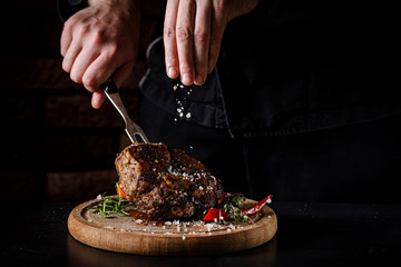 The concept of cooking meat. The chef cook salt on the cooked steak on a black background, a place under the logo for the restaurant menu. food background image, copy space text - obrazy, fototapety, plakaty