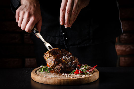 The Concept Of Cooking Meat. The Chef Cook Salt On The Cooked Steak On A Black Background, A Place Under The Logo For The Restaurant Menu. Food Background Image, Copy Space Text