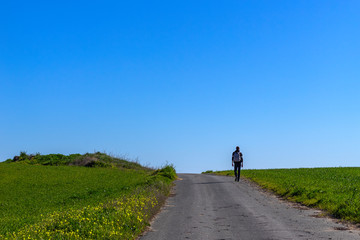Obraz premium A lonely backpacker tourist walks the road through a large field covered with green grass.