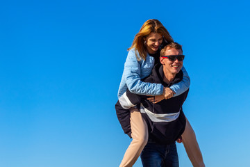 Happy man carrying his girlfriend on the back on blue sky background. Young couple enjoying travel outdoors. Love story of attractive, cheerful, funny couple.
