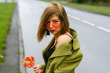 young european girl walks in rainy weather down the street with puddles of green cloak and eats...
