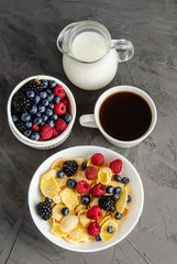 Healthy breakfast with cornflakes in a white plate, berries, milk and a cup of black coffee on a dark gray background.