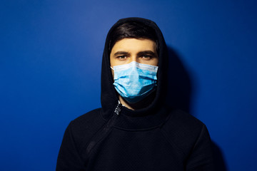 Studio portrait of young man wearing medical flu mask and hoodie sweater on background of phantom blue color.