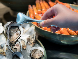 Hand of Asian woman pick a tasty fresh shucked oyster on ice with tongs. Raw oysters packed with...
