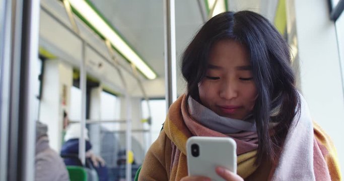 Young pretty Asian girl typing and texing on the smartphone in the tram while going somewhere and looking at the window.