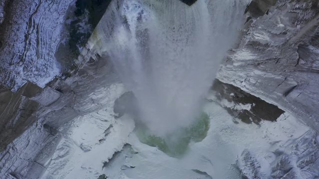 Aerial 4K footage directly above tall waterfall dropping over rocky cliff in winter. Camera looking straight down with drone rising up.