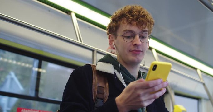 Young red-haird Caucasian guy in glasses and headphones tapping and texting on the smartphone while listening to te music and going somewhere in the tram or bus.