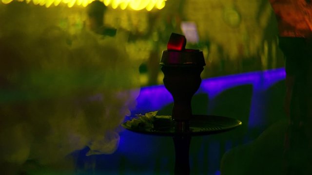 Close-up of hookah coals and smoke on background of bar. Stock footage. Hot coals of hookah with thin smoke on blurred background of bar's semi-darkness in neon light