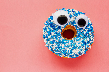Vanilla Dip Donut with Black Wiggle Googly Eyeballs on a Coloured blue and white Sprinkles on a pink background.