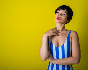 Brunette in a striped swimsuit posing in the studio on a bright yellow background. Woman in a short wig with sensual lips.