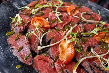 Fototapeta na wymiar Closeup Beef carpaccio with pesto sauce and tomatoes on black table. Restaurant menu, natural and organic food concept.