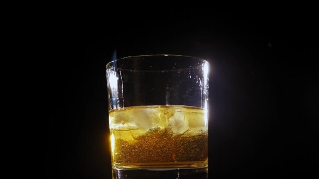 Close-up of ice cubes falling into a rotating glass of whiskey or cognac on a black background, a bright light illuminates the glass creating Golden highlights. Slow motion.
