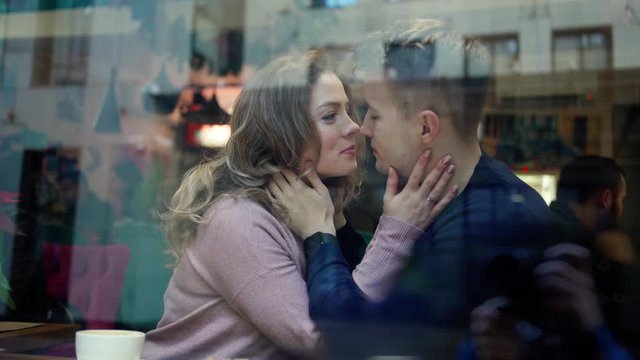 cute Happy romantic couple embracing and kissing while sitting over the window in cafe. young man tenderly hugging and kissing girlfriend in cozy cafe. View through cafe window.