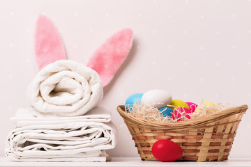 A close-up of a stack of clean white bedding, towels and Easter bunny ears, a basket of eggs on a...