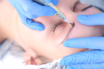 Cosmetologist making eyebrows microblading procedure in beauty salon for girl using tattoo apparatus, eyebrow closeup. Beautician in gloves is doing permanent makeup to woman. Beauty industry concept.