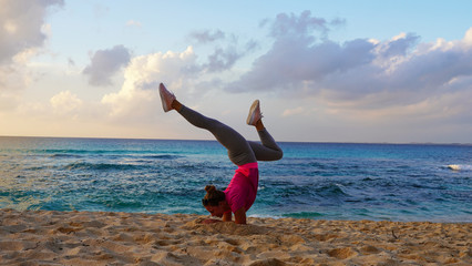  Beautiful bright girl does yoga on the ocean.Yoga on the beach, flexible sports model performs gymnastic elements, handstand and gymnastic bridge on a background of sunset blue ocean
