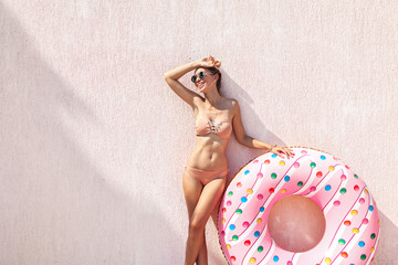 Slim girl in bikini with inflatable donut posing against pink wall - obrazy, fototapety, plakaty