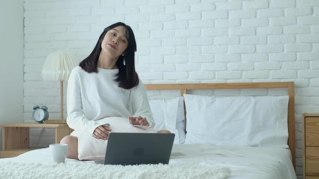 A young woman streaching neck and back while using laptop on bed, study and work on computer, modern technology