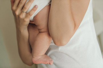 mother in white clothes hugs her little newborn baby with bare chubby legs with folds very tightly and reverently on a plain beige background