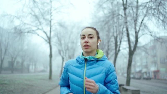 slow motion young sporty woman running with Wireless Earphones. slow motion young sporty woman in blue down jacket running along the alley at foggy winter day and uses Wireless Tws Earphones
