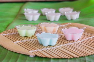 Colorful of Thai coconut jelly in small plastic cup on wooden plate with green banana leave background. Thai dessert.