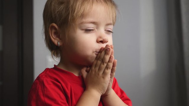 A blonde girl prays in her room alone. The child holds his hands together.