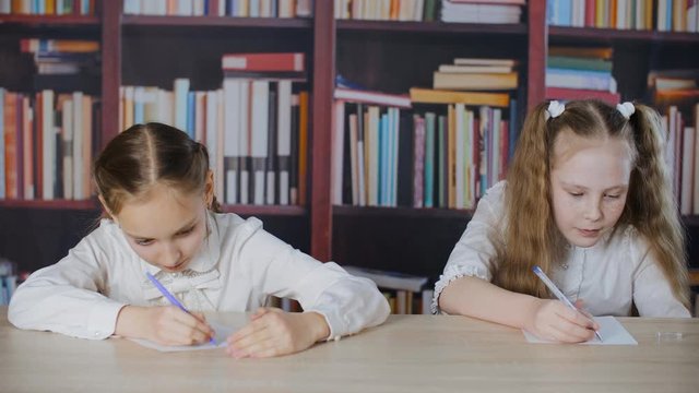 Schoolgirls writing from cheat sheet on school exam at bookcase background. Two student girl solving task with cheat sheet at school test in class on bookshelf background