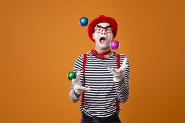 Mime in vest and red hat juggles with colorful balls on orange background - obrazy, fototapety, plakaty