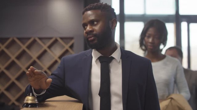Tracking with slowmo of annoyed black man in suit standing at hotel reception desk and ringing bell, other guests queuing behind him