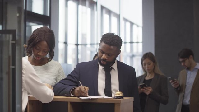 Slowmo tracking of African-American man and woman registering at hotel reception desk with help of unrecognizable female receptionist