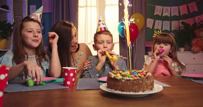 Cheerful and happy Caucasian kids with birthday boy and mother sititng at the table with a cake and celebrating birthday.