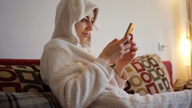cheerful smiling young woman in funny bunny pajamas sitting on red sofa using smartphone, woman looking, texting and smiling
