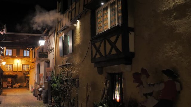 Callej&oacute;n de noche con fachadas de madera entramada saliendo humo de los hogares