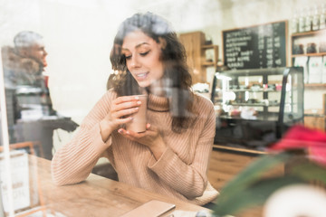 Girl holds a paper cup with coffee. Young beautiful girl is looking through the window. Girl with a cup of coffee in her hands, closed her eyes and enjoys the aroma, behind the glass in a cafe.