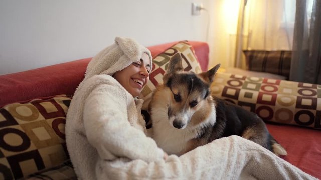 happy smiling woman hugging cute Welsh Corgi dog lying on sofa at home. Beautiful girl in funny bunny pajamas hugging funny puppy, laughing, enjoying time together, has good relationships with pet