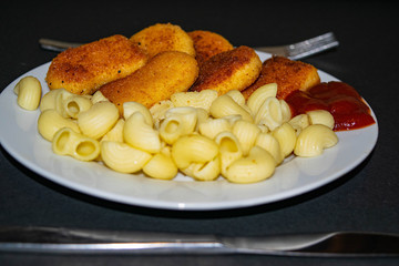 Fried chicken nuggets and boiled pasta with ketchup on a white plate on a dark background. Close up.