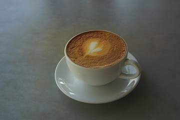 Tasty drinking, a cup of capuccino coffee decorated with heart pattern on brown milk froth in white ceramic cup on gray table