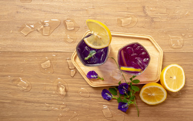 Two glasses of Blue and Violet Butterfly pea flower juice drinking, decoreted with yellow lemon sliced and fresh Asian pigeon wings flowers, on wooden table with ice, top view image