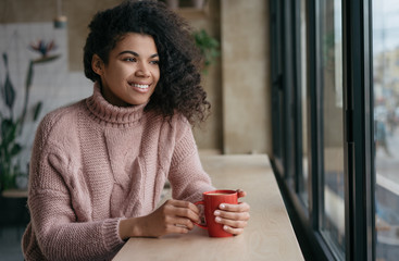 Happy African American woman drinking coffee in modern cafe. Young beautiful girl holding cup of tea, looking at window and smiling