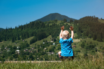 Little boy sits on mountains background with hands raised up. Back view