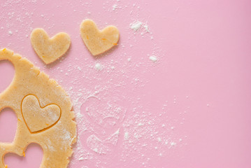 Cutting out heart-shaped cookies from flour-flavored dough on a light pink surface. Top view, the process of preparing homemade treats for Valentine's Day.