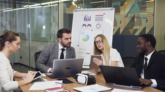 Charming blond businesswoman get aquainted with the datas on her tablet pc and on flip chart presentation to all present multiracial considerate business partners